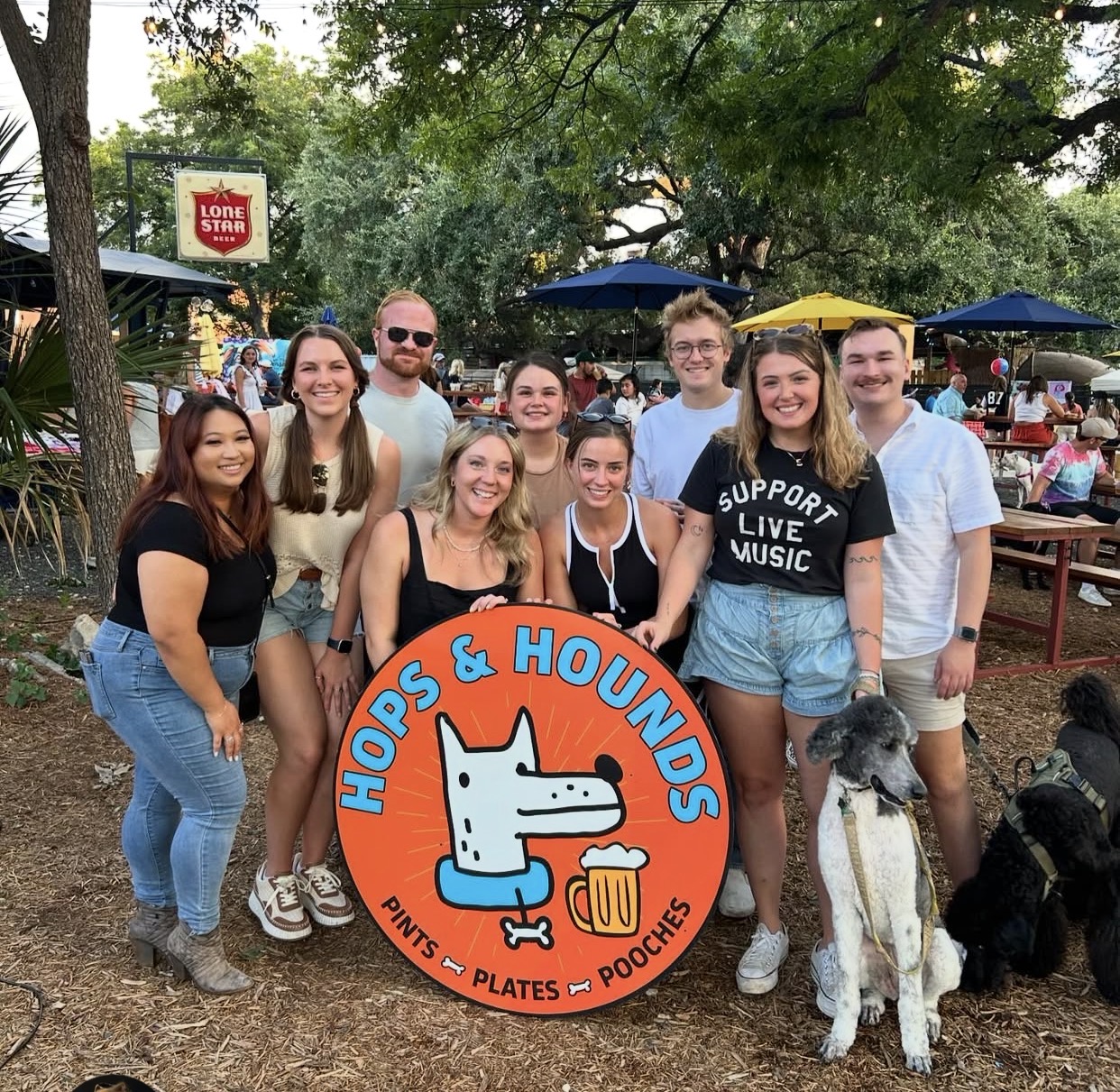 A group of friends and their dogs holding the Hops & Hounds sign at the park